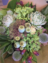 A bouquet of assorted flowers and greenery including succulents and seed heads, with a mix of green, blue, and pink colors.